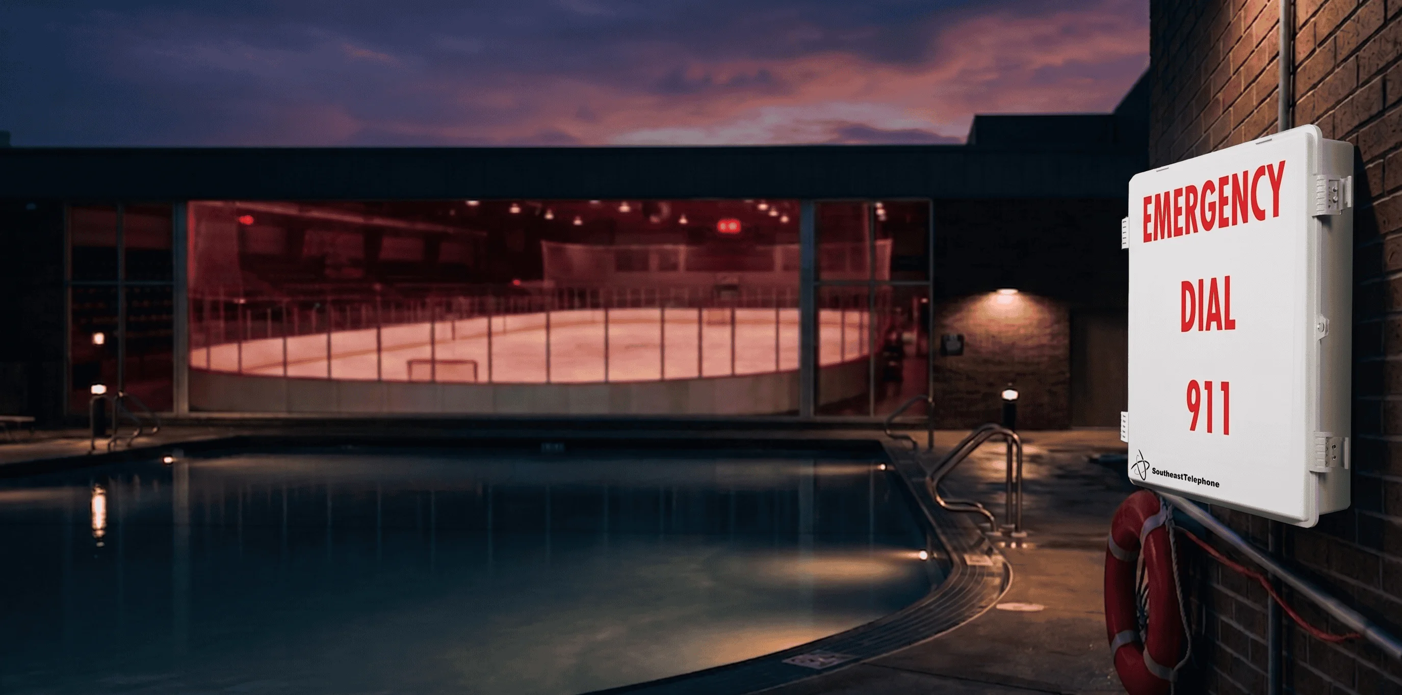 Community pool at dusk with hockey rink visible through windows and emergency phone on brick wall